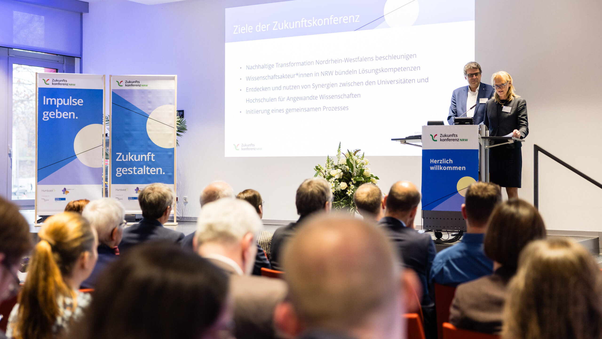 Prof. Brigitta Wolff und Prof. Oliver Locker-Grütjen auf der Bühne der Eröffnungsfeier. Publikum folgt den Vorträgen. 