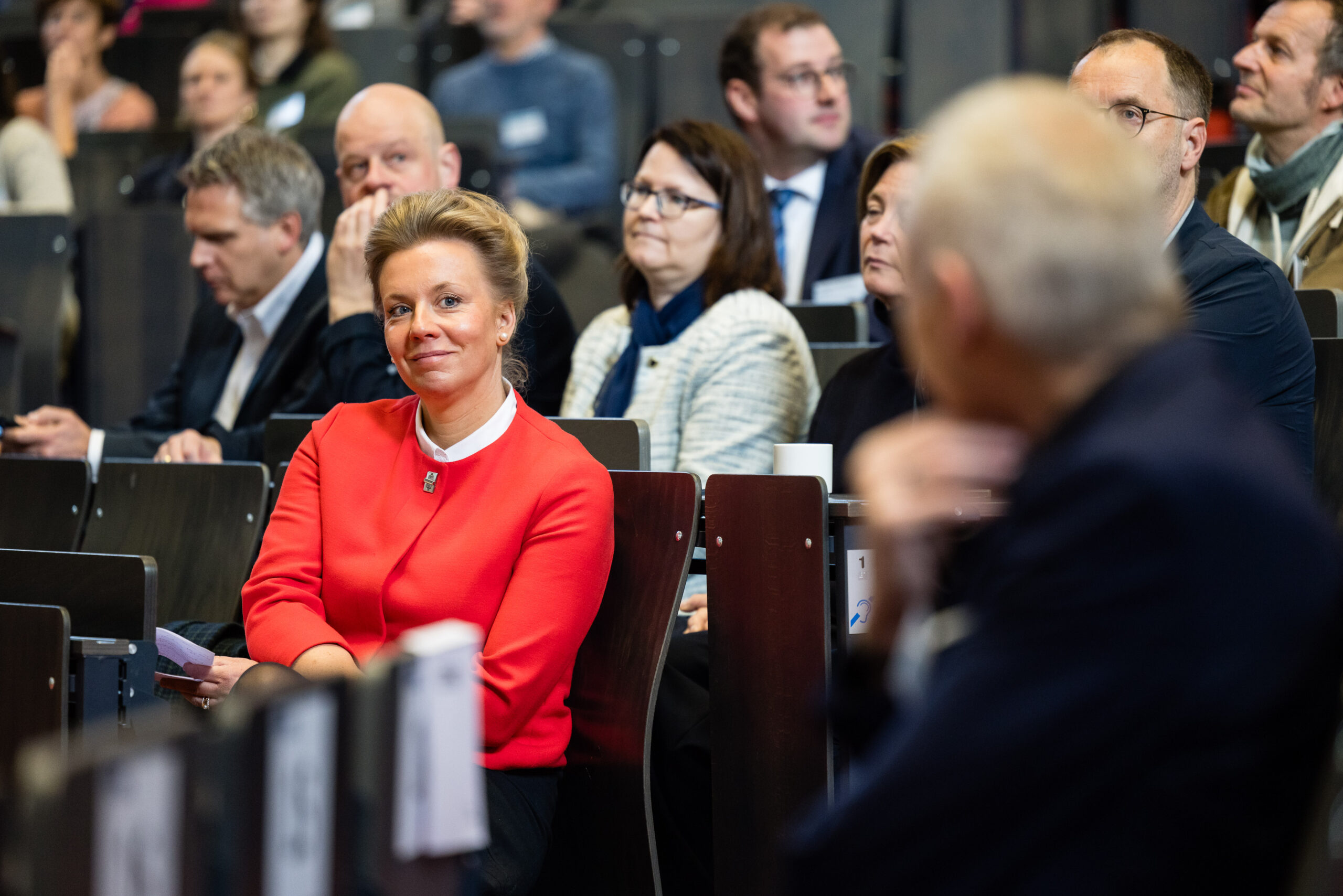 Frau mit zurückgesteckten blonden Haaren und rotem Blazer (Ministerin Ina Brandes) sitzt im Hörsaal und blickt lächelnd Richtung Kamera. Um sie herum sitzen weitere Menschen.