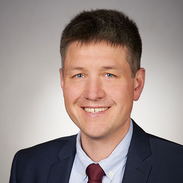 Porträtbild eines Mannes mit kurzen, braunen Haaren und blauen Augen vor einem grauen Hintergrund. Er trägt ein hellblaues Hemd mit weinroter Krawatte und einem dunkelblauen Sakko und lächelt freundlich in die Kamera.