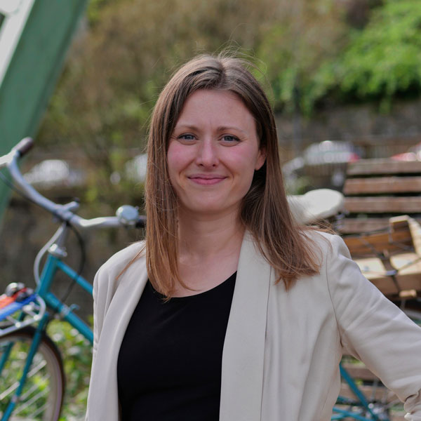 Univ.-Prof. Dr.-Ing. Heather Kaths Ein Porträtbild einer Frau mit langen, glatten, hellbraunen Haaren, die freundlich in die Kamera lächelt. Sie trägt ein schwarzes Oberteil und einen beigen Blazer. Im Hintergrund befindet sich ein Fahrrad und Natur.
