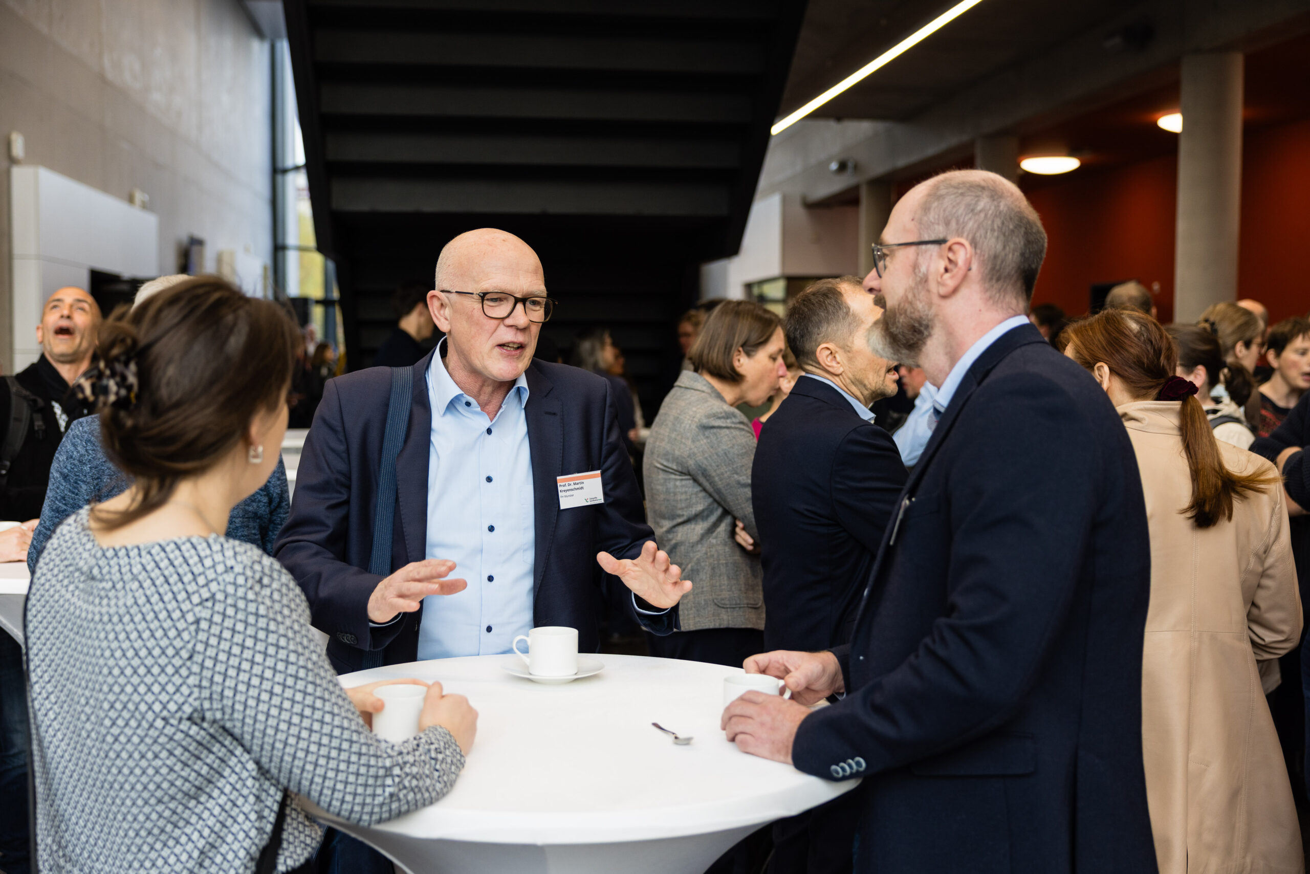 Ein Gespräch zwischen drei Personen an einem runden Stehtisch im Foyer während einer Veranstaltung; im Hintergrund sind viele weitere Persoinen zu sehen die sich unterhalten.
