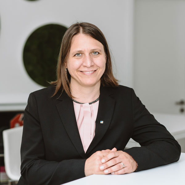 Porträtbild einer Frau mit glatten, hellbraunen Haaren und blauen Augen. Sie trägt eine rosa Bluse und einen schwarzen Blazer. Sie sitzt an einem Tisch, hat die Hände in einander verschränkt auf dem Tisch abgelegt und lächelt freundlich in die Kamera.