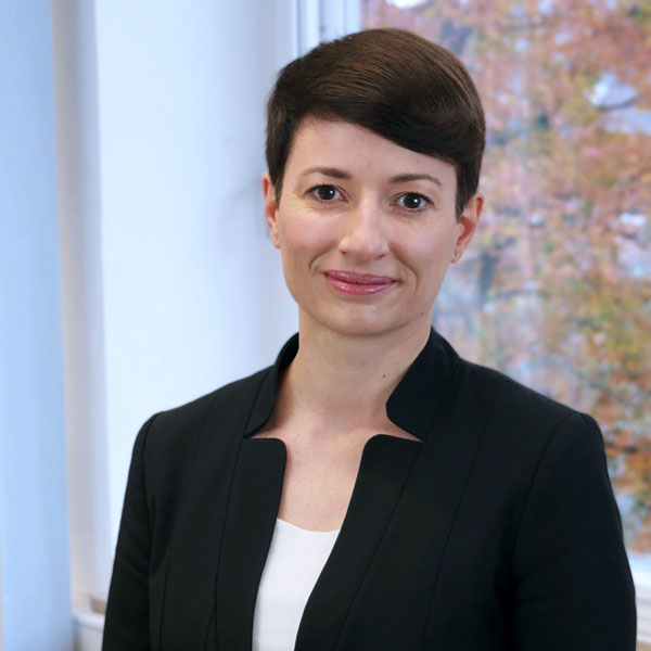 Prof. Dr. Helena Stehle | © Nadine Daum, IfK Ein Porträtbild einer Frau mit kurzen, dunkelbraunen Haaren und braunen Augen, die in die Kamera lächelt. Sie steht vor einem Fenster mit Blick auf einen Laubbaum und trägt einen schwarzen Blazer.